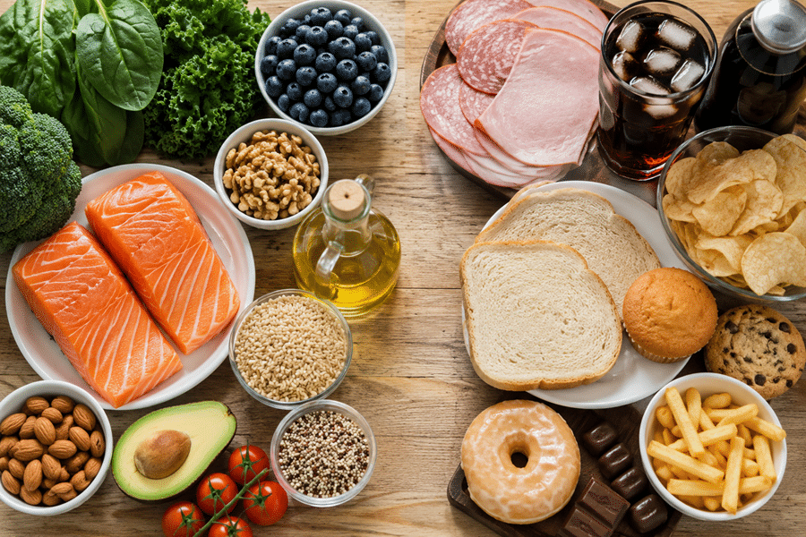 A top-down spread of fresh and prepared foods including salmon, avocado, leafy greens, berries, nuts, bread, chips, donuts, chocolates, and beverages on a wooden table.